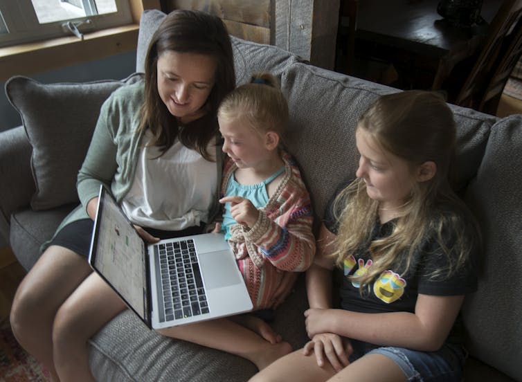 A woman and two young girls sit on a couch smiling as they look at the screen of a laptop.