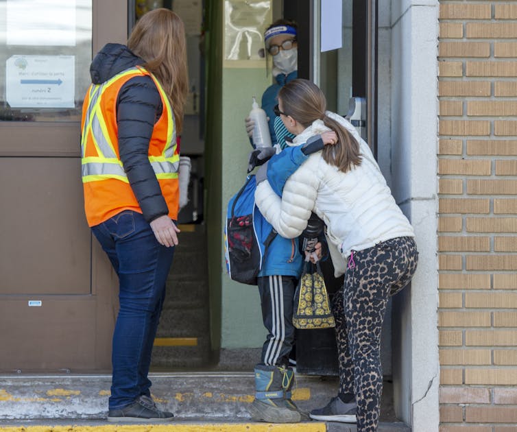 A mother and son hug as the boy makes his way into his school as a person in an orange safety vest looks on.