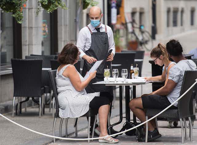 A waiter wearing a face mask takes orders from three diners on an outdoor patio.