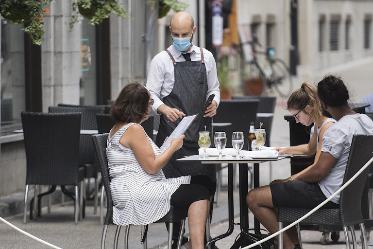 A waiter wearing a face mask takes orders from three diners on an outdoor patio.