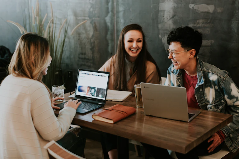 Three people sit at a table socializing with laptops and notebooks.