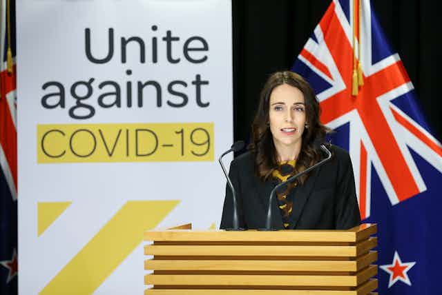 Jacinda Ardern at a lectern with NZ flag in background