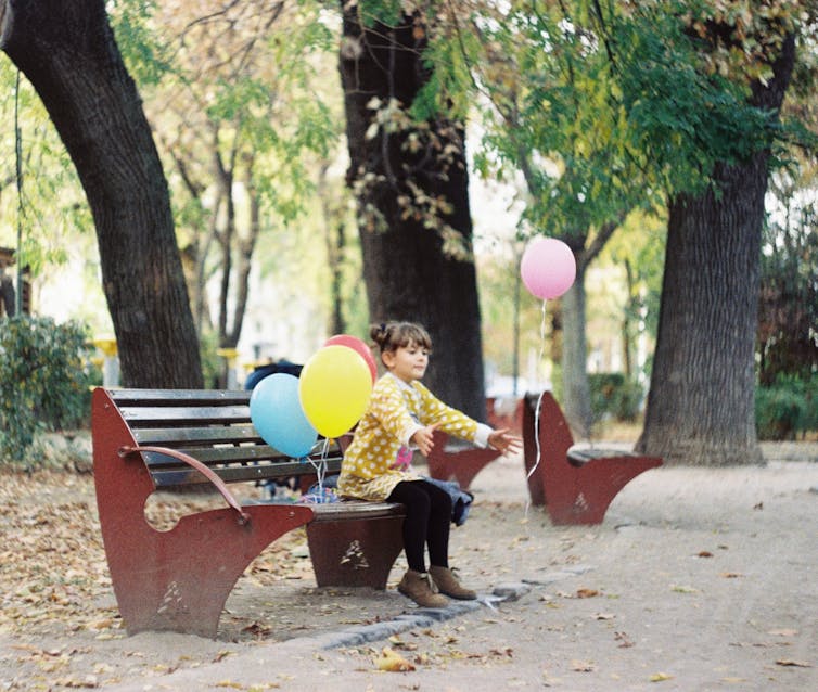 A little girl on a park bench lets go of a pink balloon