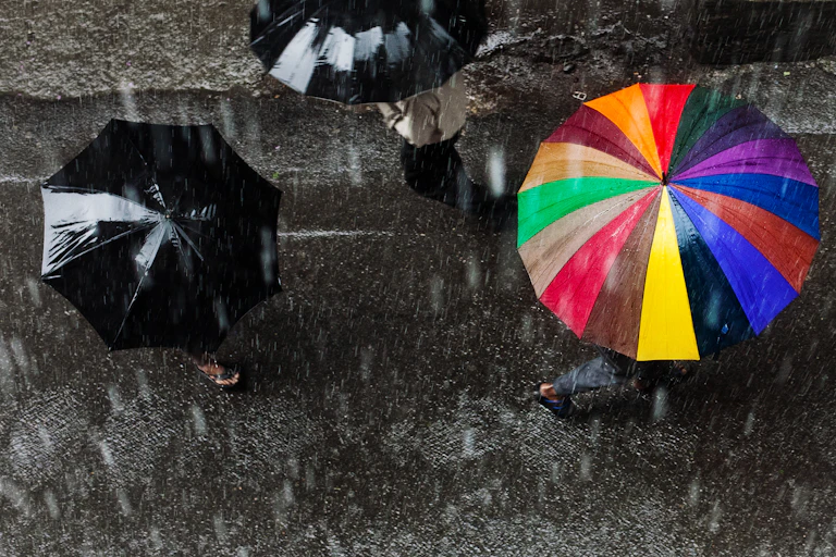 View from above of three people holding umbrellas in a heavy rain, one umbrella is rainbow colored and two are black.