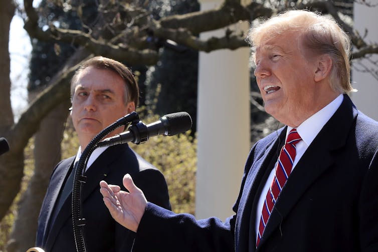 Trump gestures while speaking into a microphone with Bolsonaro looking on.