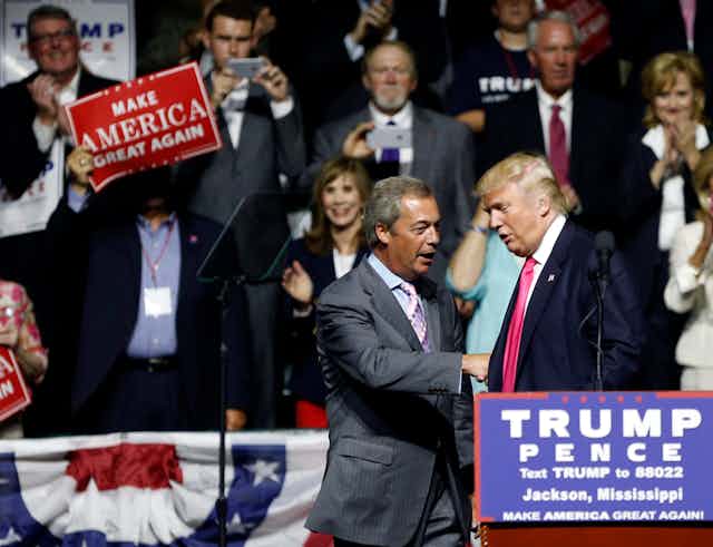 Farage and Trump shake hands in front of a Trump-Pence podium as someone waves a Make America Great Again sign.