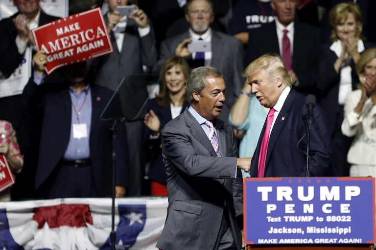 Farage and Trump shake hands in front of a Trump-Pence podium as someone waves a Make America Great Again sign.
