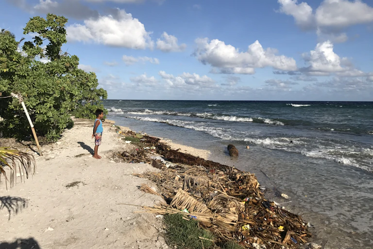 A boy with his hands behind his back stands on the sand looking out at the beach.