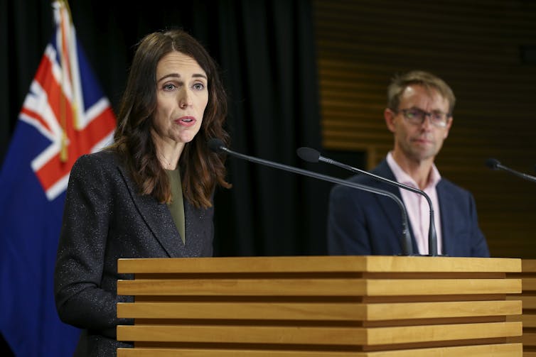 Jacinda Ardern at a lectern with NZ flag in background