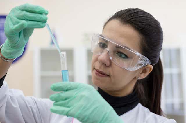 A woman wearing goggles and lab coat holding a test tube and pipette.