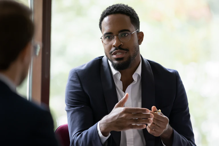 A Black man sits in an office talking to a white man, who is in the foreground with his back turned to the camera.