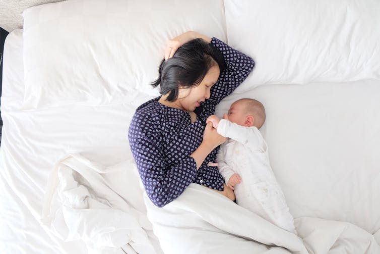 A woman lies in a bed with white bedding cuddling her baby.