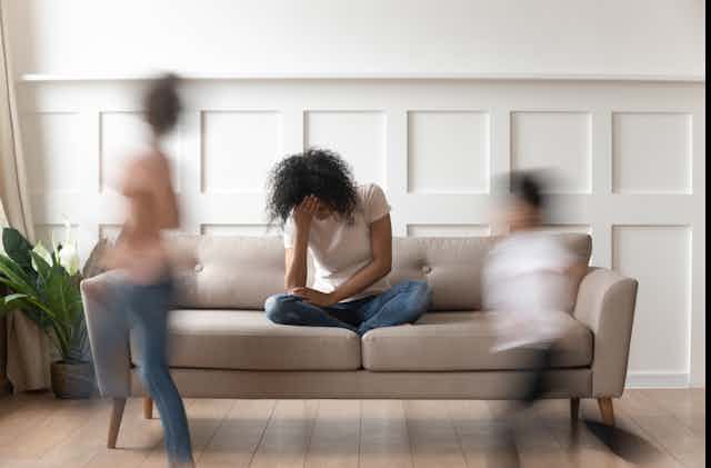 A woman sits on a sofa with her head bowed while two out-of-focus children run past her