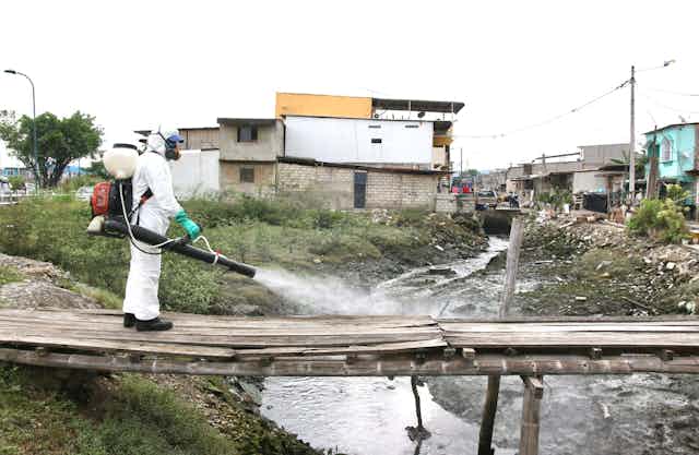 Man in protective suit sprays disinfectant over a makeshift bridge across a sewage flow in a settlement in Ecuador.
