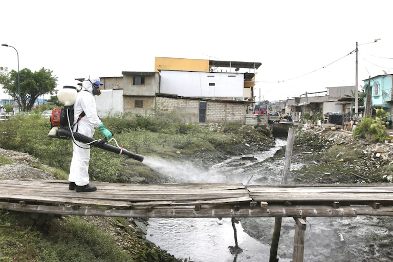 Man in protective suit sprays disinfectant over a makeshift bridge across a sewage flow in a settlement in Ecuador.