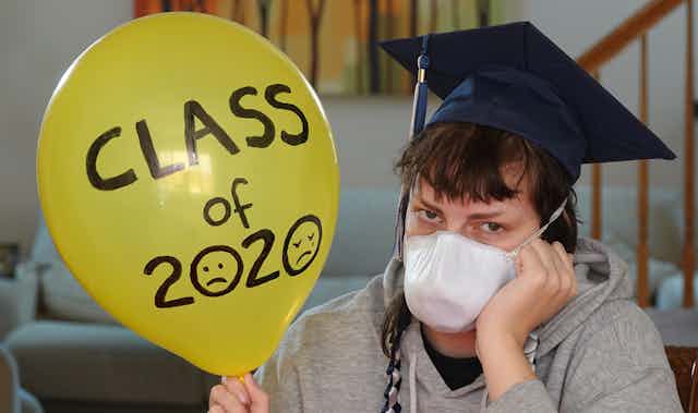 Depressed student with graduation hat and mask sitting at home holding balloon with "class of 2020" written on it.
