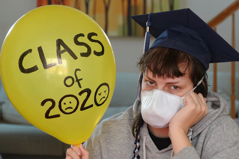 Depressed student with graduation hat and mask sitting at home holding balloon with "class of 2020" written on it.