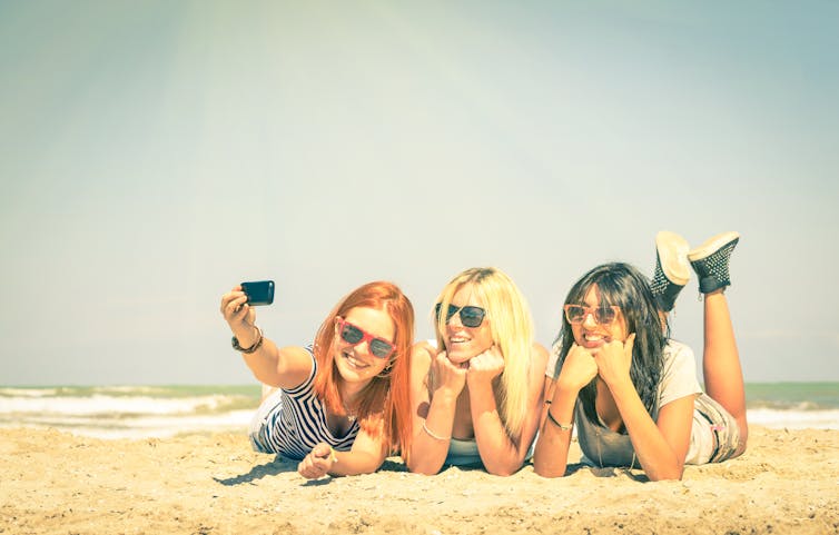 Three girls lying on towels on the beach and taking a selfie.