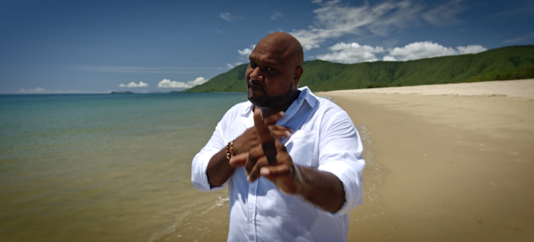 Man stands of beach, points at camera.