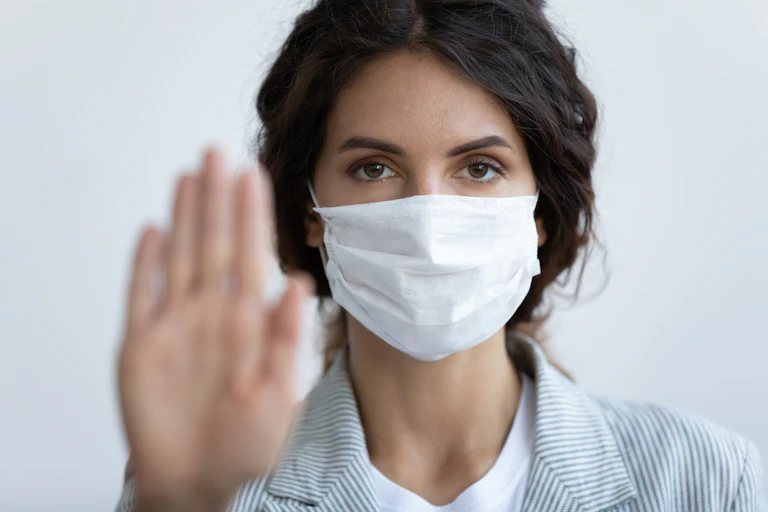 Woman wearing a mask and holding a blurred palm up to the camera, to say stop.