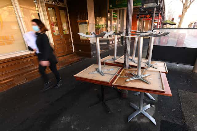 A woman, wearing a mask, walks passed some stacked tables on a Melbourne street