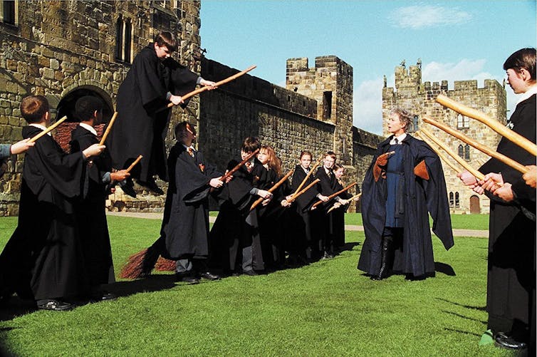 Young people dressed in black robes attempt to ride broomsticks in Harry Potter film.