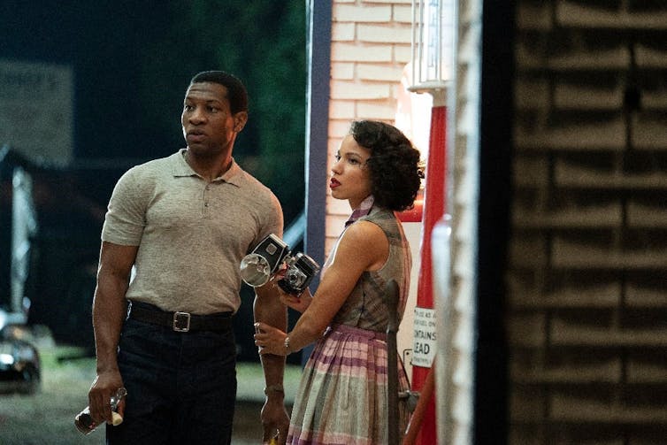 Man and woman stood outside building at night look offscreen.