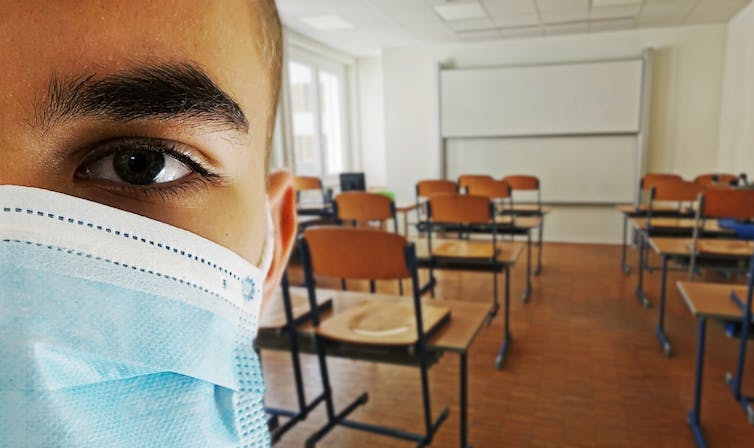 The left side of a man's masked face on the right in the foreground, with an empty classroom in the background.