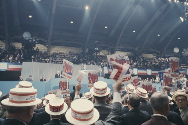 A crowd of people in straw hats wave signs at a stage.