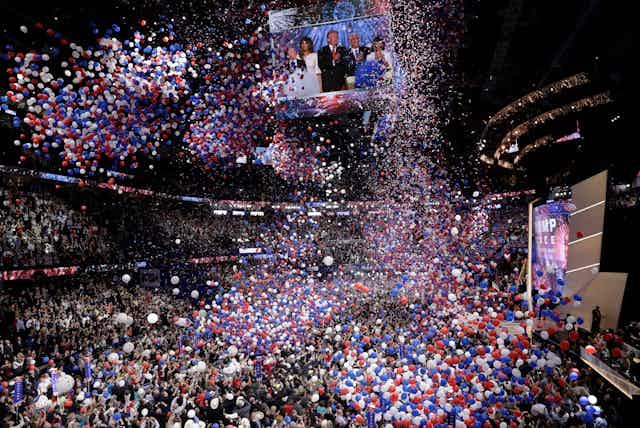 Massive numbers of balloons fall from a convention center ceiling over a crowd of people.