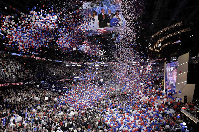 Massive numbers of balloons fall from a convention center ceiling over a crowd of people.