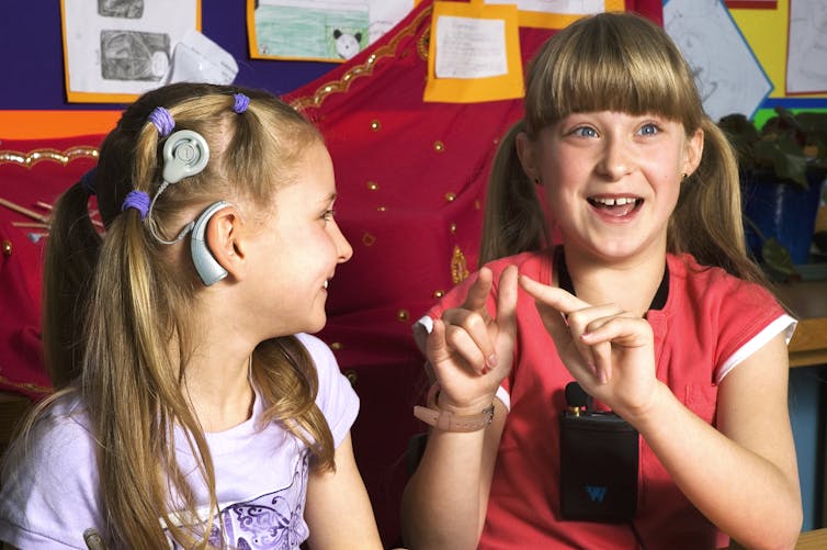 A twelve year old girl dressed in a red top and an nine year old girl dressed in a lilac top communicate with sign language. The younger girl is fitted with a cochlear implant and the older girl wears a voice amplifier.