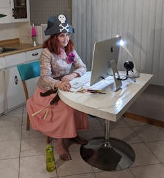 A woman in a pirate hat with pink hair sits in front of a desktop computer in a kitchen.