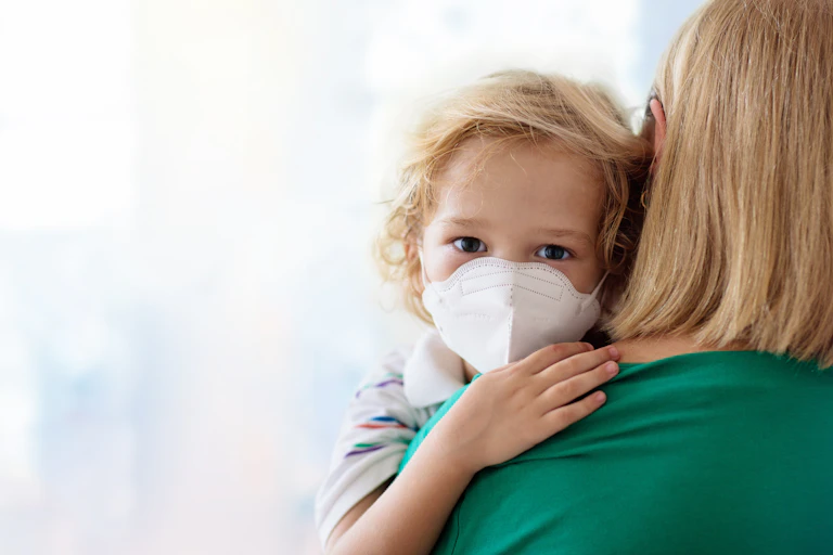 Child with face mask on being held by his mother.