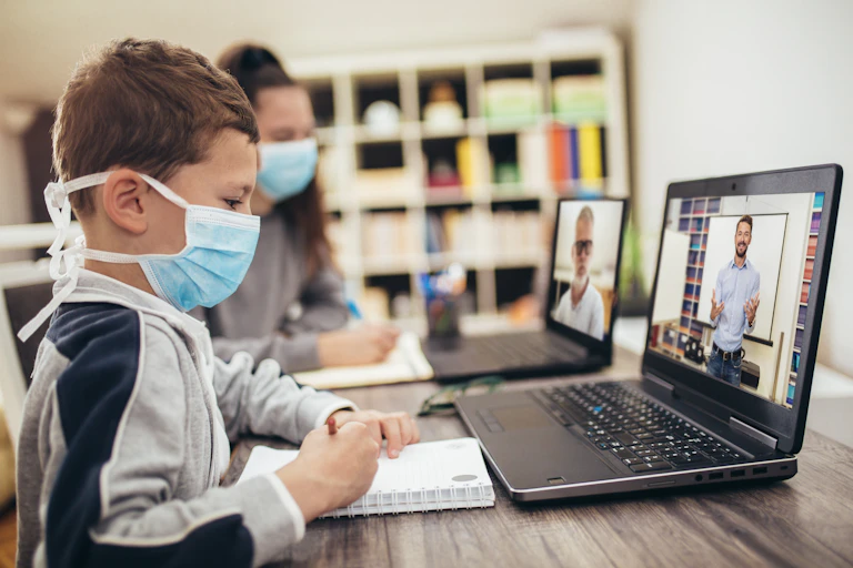 Niños con mascarilla, ante un cuaderno y un ordenador en el que se ve al profesor.