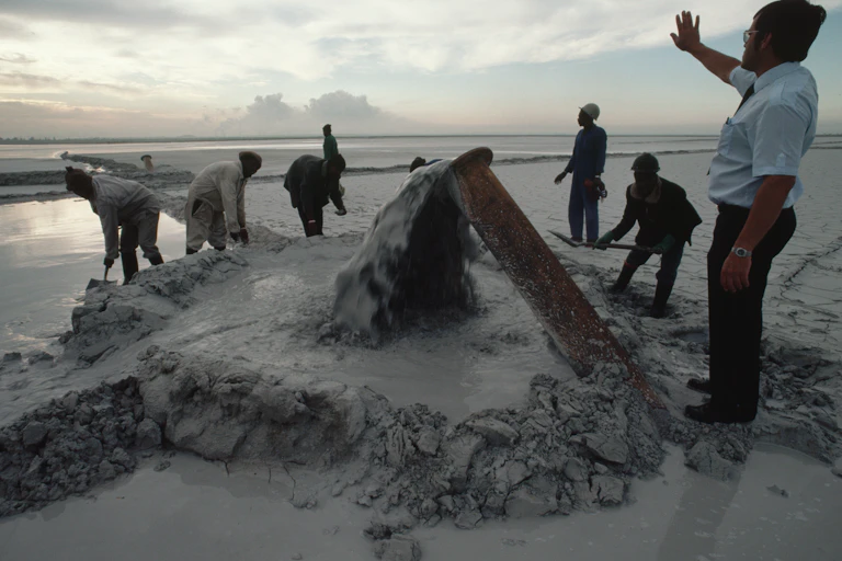 A white man holds up his hand as he instructs labourers who are digging sludge with spades as water gushes from a pipe.