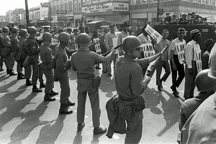 A photo from 1968 showing striking sanitation workers marching past Tennessee National Guard troops with bayonets in Memphis, Tenn.