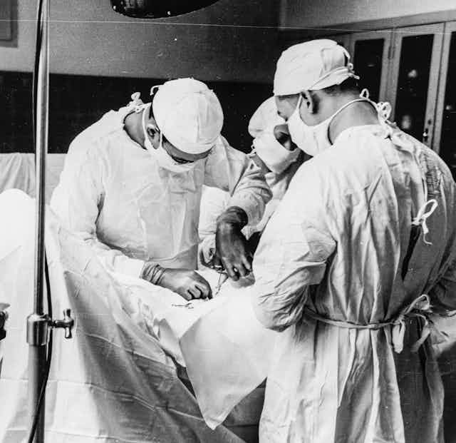 Three doctors hunched over a patient performing surgery, faces covered by surgical masks.