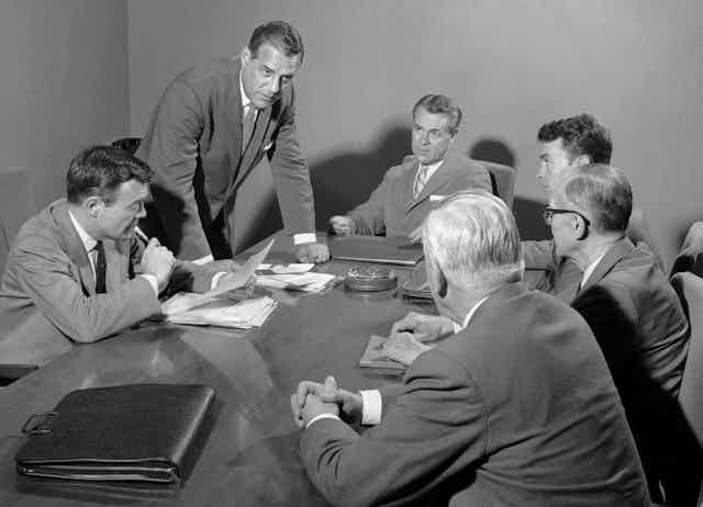 six white men in suits around a 1950s boardroom table