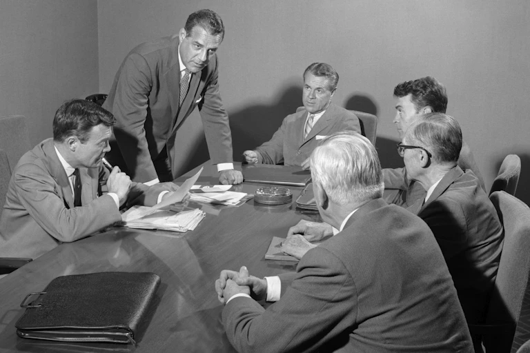 six white men in suits around a 1950s boardroom table