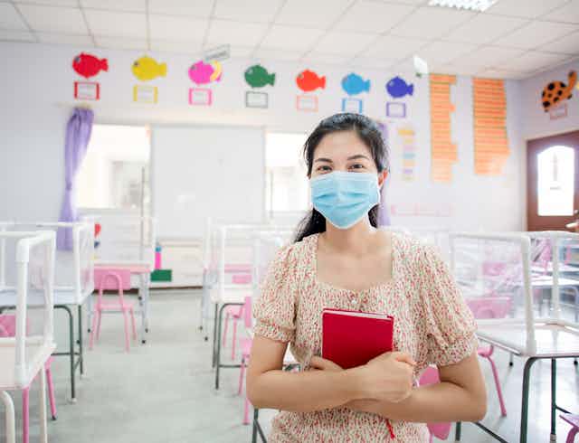 A woman wearing a blue face mask sits in an empty children's classroom, holding a red book.