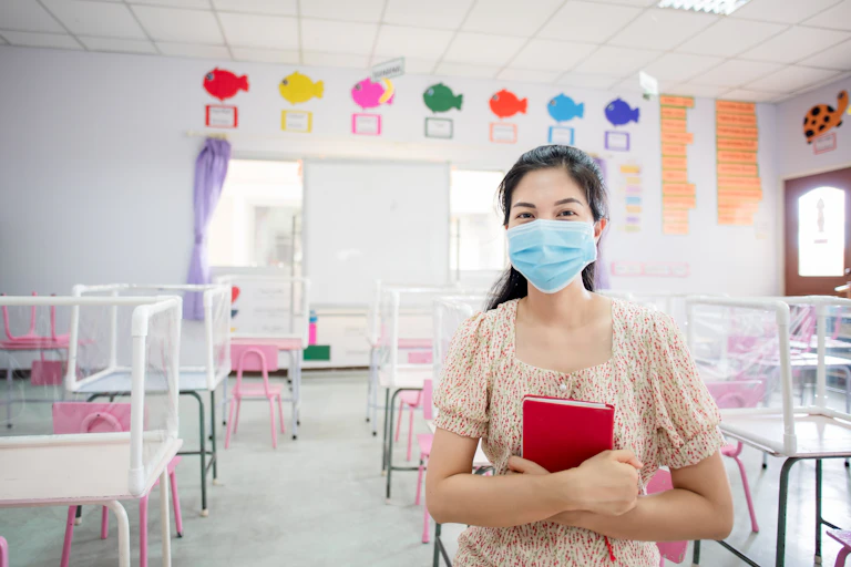 A woman wearing a blue face mask sits in an empty children's classroom, holding a red book.