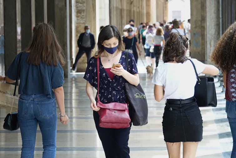 Woman wearing face mask walking on busy street checks phone