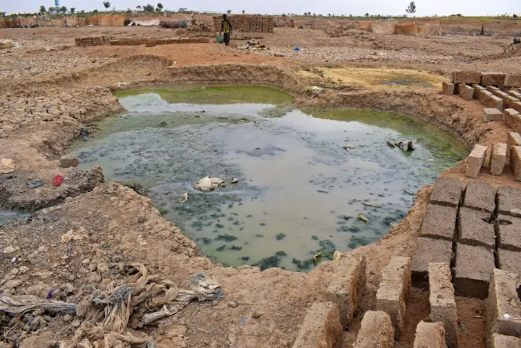 A dug-out hole with water in it and mud bricks stacked next to it.