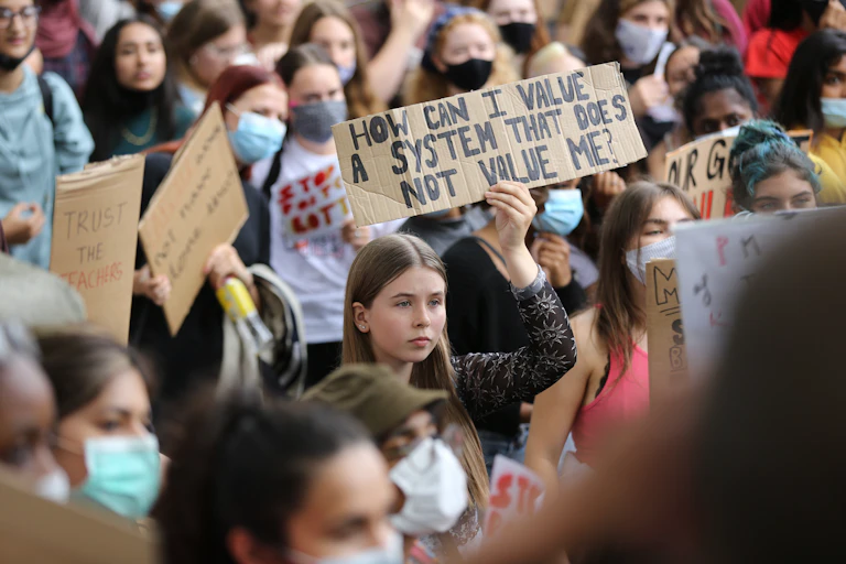 Crowd of protesting students. One holds a sign saying "How can I value a system that does not value me?"