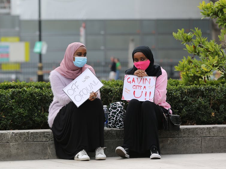 Two young female protesters sit with signs targeting Ofqual and Gavin Williamson.