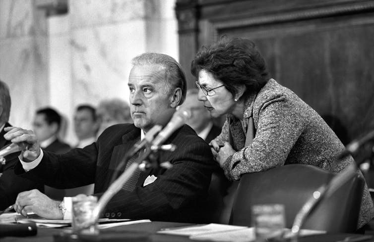 Black and white photo of Barbara Boxer speaking into a seated Joe Biden's ear in the Senate