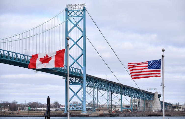 A Canadian flag and an American flag in the foreground with a suspension bridge over a river behind them.