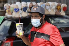 A man in a red shirt holding a soda while wearing a mask in front of a display of mannequins all wearing masks