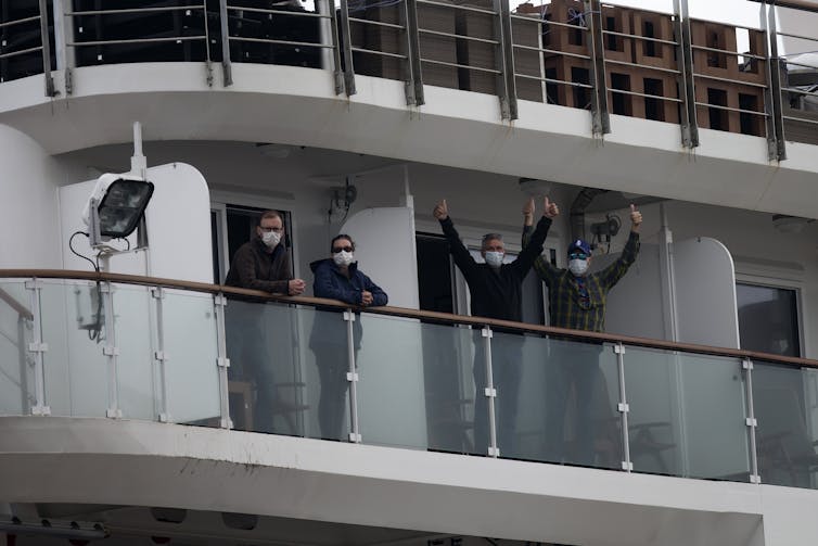 Quatre passagers masqués font signe depuis un balcon, à bord du bateau de croisière Greg Mortimer.